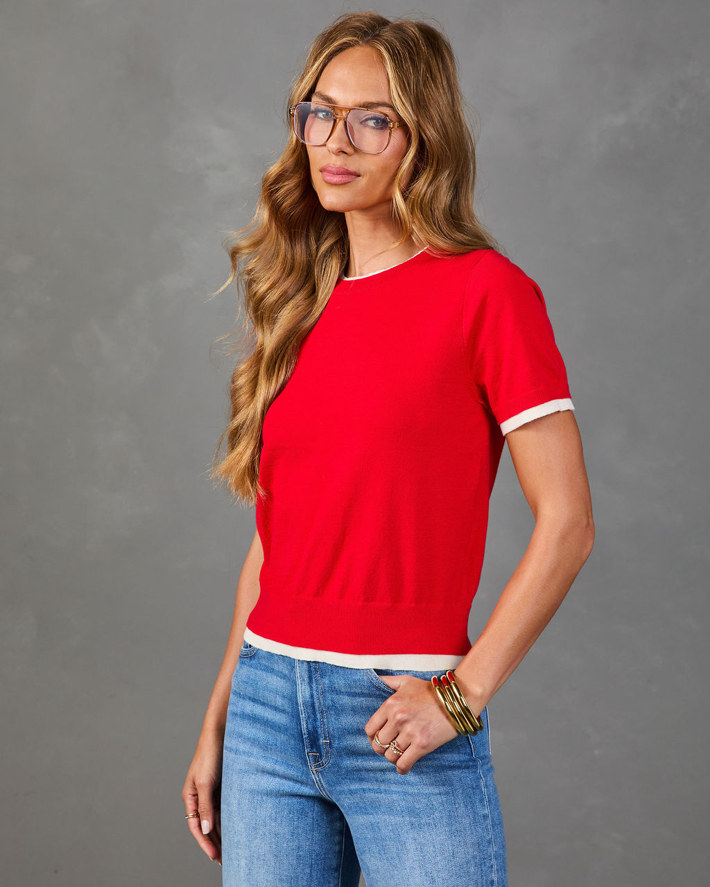 Woman wearing red short-sleeve cropped knit sweater with white trim, blue jeans, tortoise glasses, and gold bangles