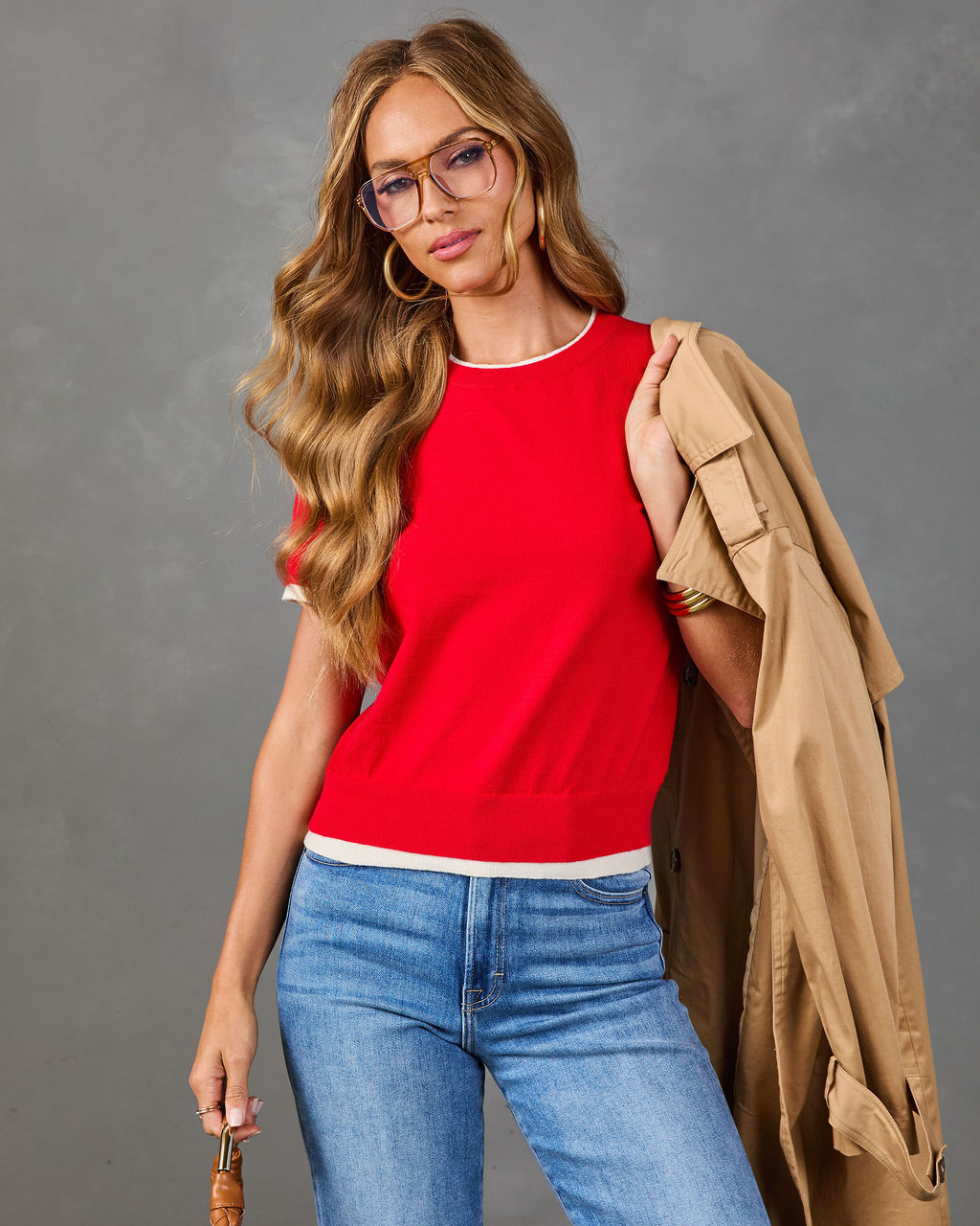 Woman wearing red knit sweater top with white trim, blue jeans, and tan trench coat over shoulder, styled with glasses