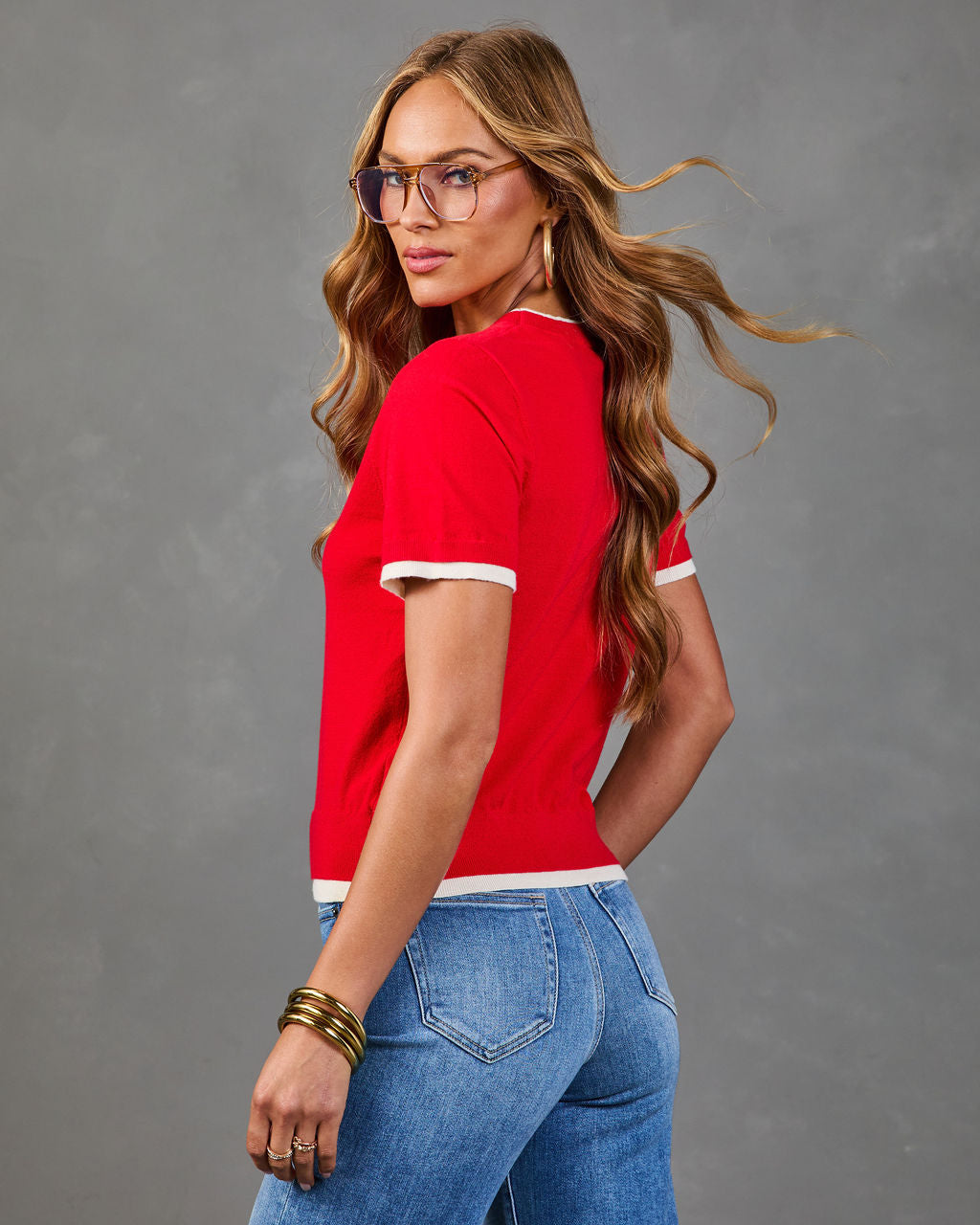 Woman in red ringer knit top with white trim, blue flare jeans, gold bangles, and tortoise glasses, posing against gray background