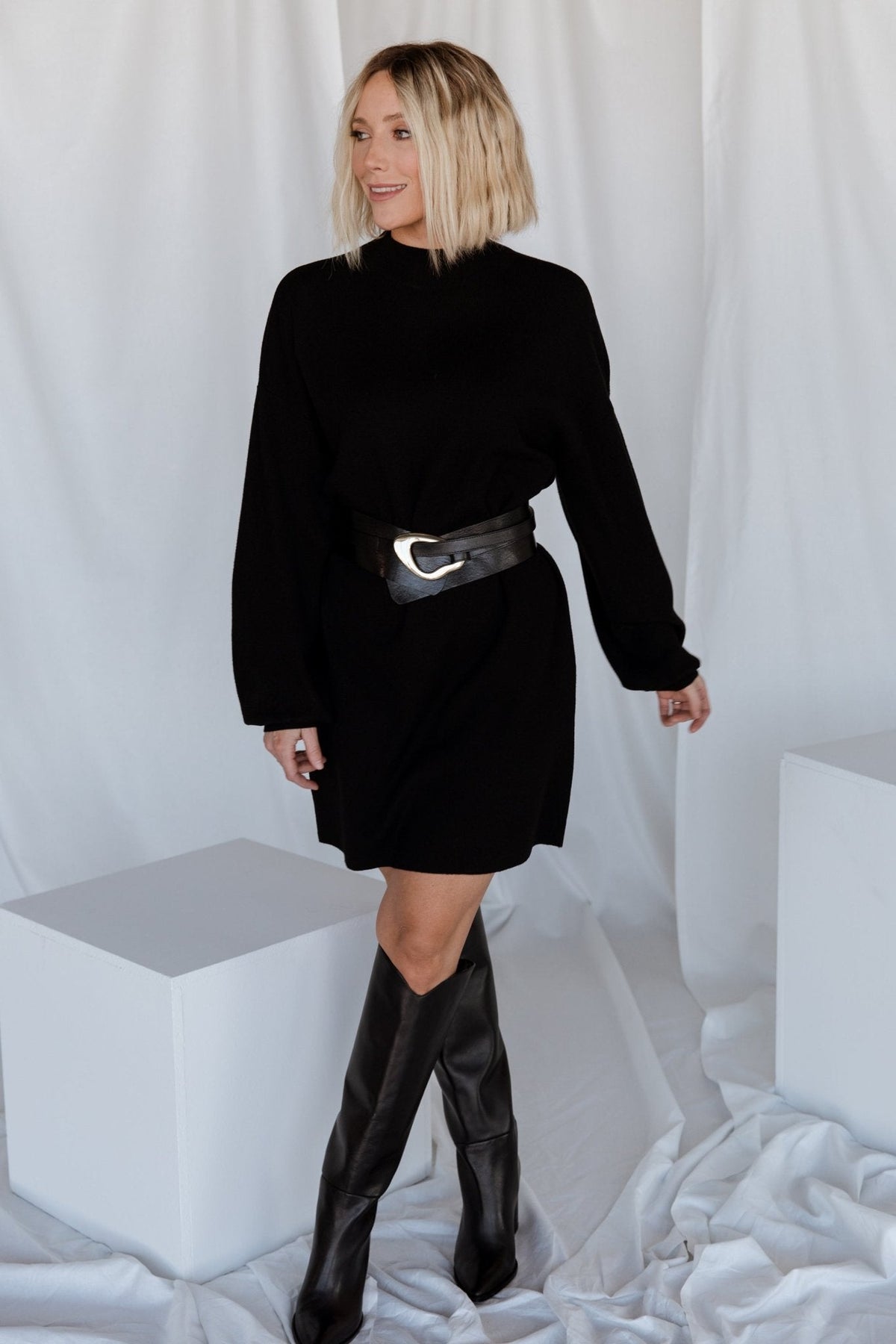 Model seated wearing the black mock-neck sweater dress, illustrating comfortable stretch and relaxed fit across the body and sleeves.